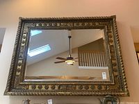 Front view of large rectangular mirror with ornate metallic frame reflecting ceiling fan and staircase.