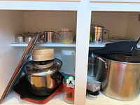 Image of lower cabinet showing large stainless steel stock pot with lid, black enamel stock pot nested inside with metal steamer basket, a metal colander, glass cutting board with red border, and various small kitchen items including a wooden steamer basket and cookie cutters.
