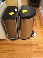 Two stainless steel trash cans side by side with black plastic lids and foot pedals, on wooden floor, in kitchen area.