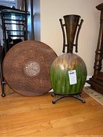 Full shot of ceramic vase with metal stand next to the round carved wooden table top.