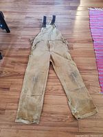Full view of the brown Carhartt work overalls laid out on hardwood floor showing general wear and dirt marks especially on legs.
