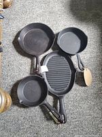 Four black cast iron cookware pieces—three round skillets and one oval grill pan with ridges—laid out on carpet.