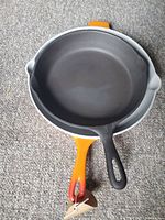 Two cast iron skillets stacked on each other on carpeted floor, smaller black one on top, larger orange enameled one underneath.