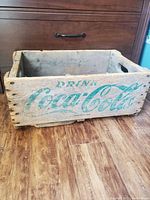 Wooden Coca-Cola crate with green logo on side, showing wear and damage.