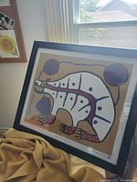 Framed colorful print of a stylized bear with distinctive Indigenous art style, placed near a window.