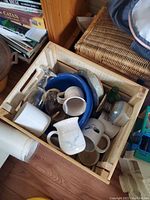 Wooden crate filled with various glass and ceramic vessels including mugs, cups, a blue bowl, and a small wooden item.