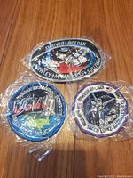 Three individually wrapped embroidered space mission patches on a wooden floor, showing detailed astronaut and spacecraft designs with mission and crew names.