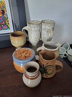 Group of seven assorted pottery items including mugs, pitchers, small vase, goblet cup and lidded jar on a wooden surface.