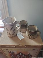 Photo of the pottery pitcher with handle next to two small cups on a box.