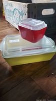 Large yellow rectangular Pyrex dish with lid and small red square dish with lid stacked on top, on wooden floor with vintage Coca-Cola crate in background