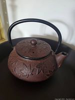 Photo of small cast iron kettle with embossed leaf pattern and fixed handle, showing body, lid, spout, and handle.
