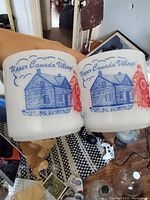 Two FireKing milk glass mugs held side by side showing Upper Canada Village blue and red graphics of a cabin and a person.