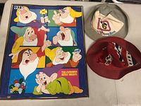 Framed Seven Dwarfs print next to two vintage Disney hats, beige cap and red bucket hat with Mickey Mouse patch