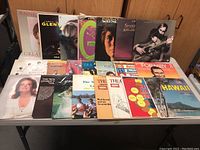 Photo showing 20 LP record albums arranged on a table with various cover images including portraits, landscapes, and group photos.