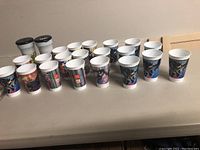 Full set of 16 white plastic McDonald's Batman cups arranged on a table showing various designs.