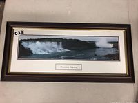 Framed panoramic photograph of Niagara Falls with a dark wood frame and white matte with gold trim, showing the falls and water.