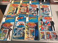 Photo showing nine sealed audio cassette sets with DC Comics themes, including Superman and Batman, featuring collector's comics and audio cassettes in blister packaging.