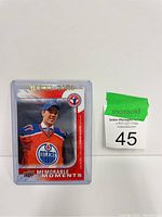 Photo of a Connor McDavid rookie hockey card in protective plastic sleeve, shown against a plain background next to a lot number tag.