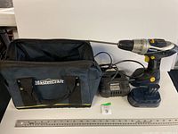 Photo showing Mastercraft cordless drill alongside its battery charger and a black soft carrying bag with Mastercraft logo, placed next to a ruler for size reference.