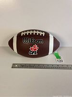 Photo of a brown Wilson CFL football with white laces and stripes, placed next to a ruler for scale showing approximately 28 cm length.