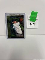Photo of Tiger Woods rookie card in a clear plastic protective case on a white background with a MaxSold auction label numbered 51 beside it.