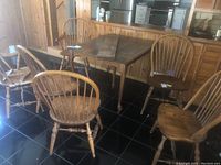 Photo showing 6 oak chairs arranged around a partial view of a dining table, including 2 armchairs with armrests and 4 side chairs without. Chairs have vertical spindles on backs and turned legs.