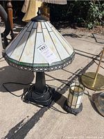 Photo showing two Tiffany style stained glass table lamps outdoors on concrete. Larger lamp has conical shade with cream/beige stained glass and green trim, smaller lamp has cylindrical shade in similar style.