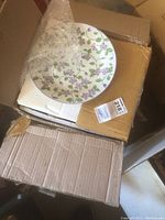 Opened cardboard box showing one porcelain plate decorated with violet flowers and green leaves, partially wrapped in bubble wrap.