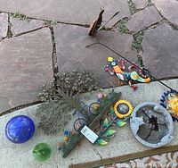 Multiple yard art pieces displayed on a stone and concrete surface including metal tree sculpture, colorful metal lizards, sunflower wall art, sun face ornament, concrete birdbath, and two glass spheres.