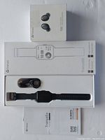 Photo of the Tranya T20 wireless earbuds box and the Tranya T-Rex smartwatch box alongside the smartwatch device, charging cable, and user manual laid out on a surface.