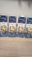Front view of four unopened gold-tone Elements by Weiser door knobs in blister packaging on a wooden surface