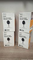 Four boxed Arenti IN1 Full HD 1080P indoor cameras stacked in two columns, front view showing product name and camera image on the boxes.