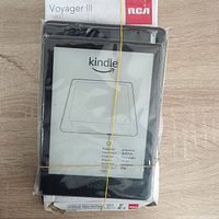 Plastic-wrapped Amazon Kindle e-reader laid on top of the RCA Voyager III tablet box and charger, secured with rubber bands