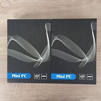 Front view of two black Mini PC boxes with design and Intel CPU, eMMC flash labels.