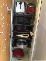 Multiple kitchen appliances and bakeware stored on a shelving unit including toasters, grills, baking pans, and mixer.