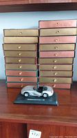 Stack of 25 boxed books with gold and pink lids, shown with silver model car in front