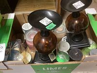 Box containing two large metal and glass candle holders, several jar candles, and pillar candles in black, green, and cream.