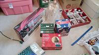 Photo showing multiple Christmas decoration boxes, porcelain lighted house figure, a white angel figurine, and a box with assorted ornaments.