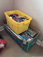 Carton filled with over 30 rolls of Christmas wrapping paper with a yellow plastic tote on top filled with ribbons and bows.