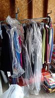 Rack of assorted clothing covered partially by clear garment bags showing multiple items including jackets, pants, dresses, and sweaters.
