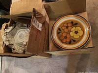Two open boxes: one contains stacked plates displaying a floral design in brown, orange, yellow hues; the other has wrapped cups and saucers inside packaging.