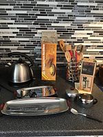 Wide view of kitchen counter showing electric kettle, loaf cake tin, cream and sugar containers, poppy seed grinder in box, cooking utensils in holder, and other kitchen items.
