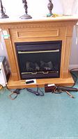 Front view of the electric fireplace with oak mantle and black insert, remote control on base, power cords visible.