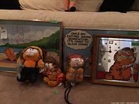 Two framed Garfield cartoon prints and three Garfield plush dolls in front of a couch, also a Garfield themed wall clock to the right.