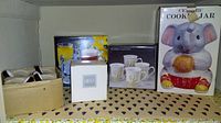 Shelf showing boxed Mikasa mug, Studio Nova Winter Rose mugs in cardboard holder, and ceramic elephant cookie jar box