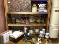 Photo showing wooden shelving with various metal and glass candle holders, a boxed Elements brand pillar candle, a basket with many white tapered candles, a decorative metal wheelbarrow with an egg, a nutcracker figurine, and red berry candle holder sets.