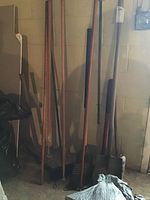 Long-handled tools leaning against basement wall including pickax, shovel, sledgehammer, axe, and brooms.