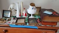 Table with large Holy Bible, coral rosary, religious figurines, framed prints, white vases, and a decorative white pedestal vase with floral design.