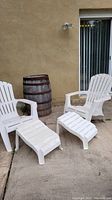 Two white plastic Adirondack chairs with two matching footrests shown outdoors, one chair visibly damaged.