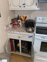 Kitchen corner view showing countertop with cooking utensils in containers, a metal tea kettle, and a black electric air fryer. Cabinet below has open doors displaying multiple pans and baking sheets stored inside.